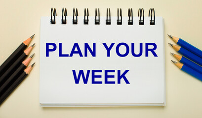 On a light background, a white notebook with the text PLAN YOUR WEEK and black and blue pencils on the sides.