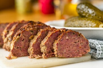 Baked sliced beef meatloaf, pickled cucumber and towel on kitchen table.