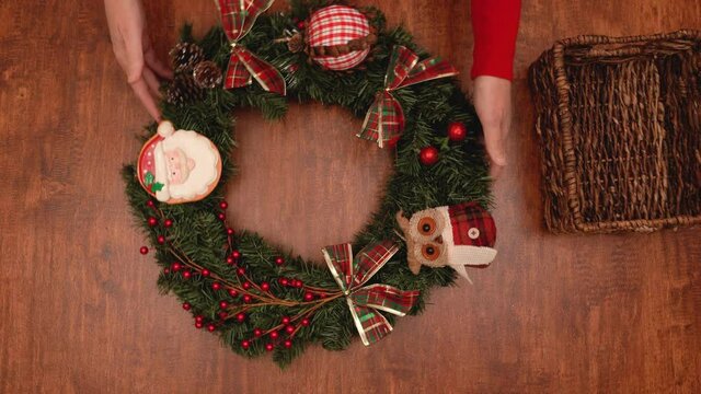 Woman decorating Christmas pine wreath with red winter berries, ornaments, and pine cones, close up view from above on wooden background