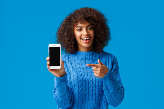 Check this out. Happy charismatic african-american woman with afro haircut, holding smartphone, showing mobile screen, pointing display as promote application, shopping app or game