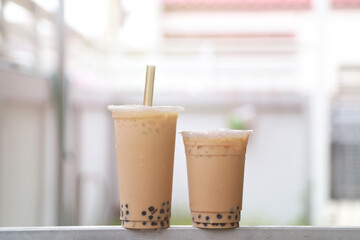 Two ice milk tea and bubble boba in big and small plastic cup put on bar steel and sun light
