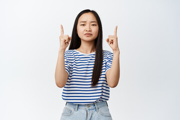 Skeptical young asian woman has doubts, frowns and looks reluctant, points fingers up at...