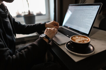 professional man with a luxury watch in a black hoodie works on a laptop and drinks coffee at a...