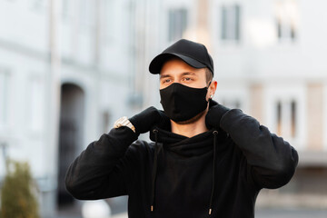 Fashionable young man with a protective medical mask in a black stylish hoodie with a fashion cap on the street. Modern male urban style and pandemic concept