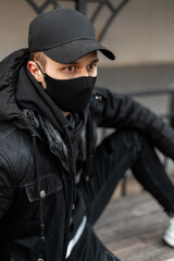 Fashionable handsome young man with protective black mask in fashion black jacket and hoodie with cap sits on the street