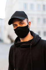 portrait of handsome young hipster man model with protective medical mask and black fashion mockup cap wearing trendy hoodie in the city
