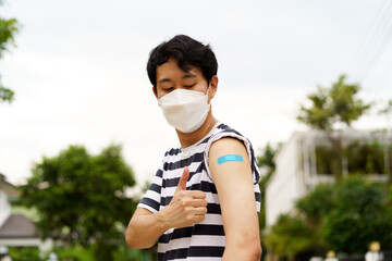 Young Asian man wearing covid-19 face protection mask showing thumbs up gesture at while looking at bandage after vaccine for protection against corona virus during day