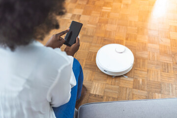 Relaxed woman with phone and robotic vacuum cleaner. Woman using automatic vacuum cleaner to clean the floor, controlling smart machine housework robot with smart phone