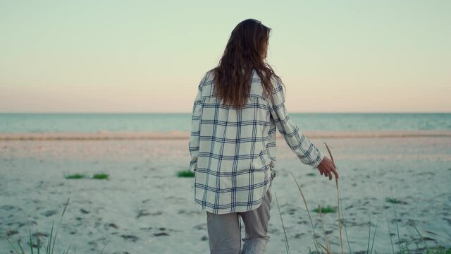 woman hipster walks on wild sea sand beach at sunset and touches spike of grass by hand. leisure outdoor vacation, escape to nature, camping lifestyle