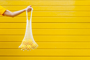 Female's strong hand holding reusable mesh bag of lemons fruit on yellow wall background. Sustainable lifestyle. Plastic free, zero waste concept.