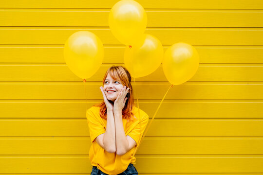 Bright picture of red haired positive female posing with yellow helium balloons on yellow background in the city street outdoor. Happy emotion caucasian woman enjoy standing over yellow wall.