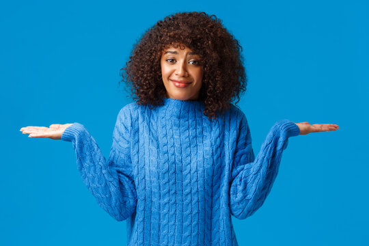 Who cares i dont know. Confused and clueless silly cute smiling african-american female in winter sweater, shrugging with hands spread sideways and smile as have no idea, standing blue background