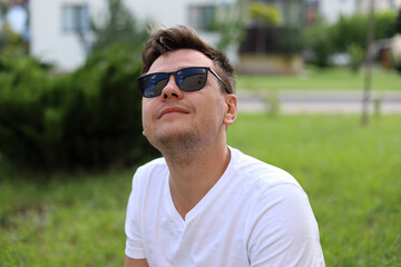 Naklejka na ściany i meble Smiling looking up young caucasian man with sunglasses in a white t-shirt on park with blurred sunny background. Selective focus. Portrait of a 32 year old young man.
