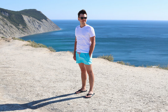 Smiling Looking Sideways Young Caucasian Man With Sunglasses In White T-shirt And Blue Shorts On Mountain On Sea Blurred Sunny Background. Portrait In Full Growth Of A 32 Year Old Young Man.