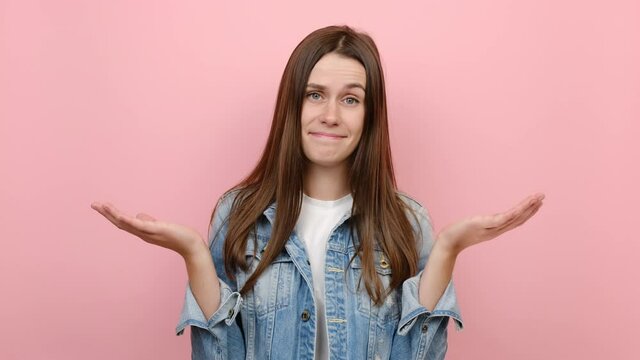 Fun shy ashamed caucasian female 20s years old look camera spreading hands say oops ouch oh omg i am so sorry, wears blue denim jacket, posing isolated on pastel pink color background in studio