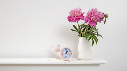 Red, pink peonies in a white vase on table, clock, and against a white wall background