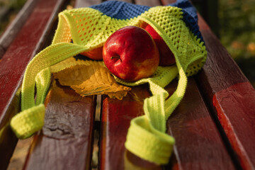 Red apples on the bench in yellow blue ecofriendly string bag on a blurred background.