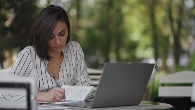 Business Woman Brunette Arabic Hispanic Ethnicity Works remotely while sitting in a summer cafe on a sunny day with a laptop and writes down with a pen and notebook
