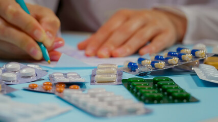 Doctor hand writing a prescription. Nearby Pile of pills in blister packs on blue background. Close Up.