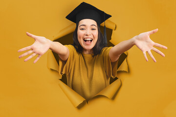 Happy asian girl, with graduation hat, comes out of a hole on a yellow paper backdrop