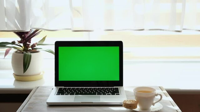 Mock-up laptop computer with chroma key green screen on coffee table houses near window.Cup of hot tea near computer.On background sunny window, flower and curtain.Concept: online shopping, freelance