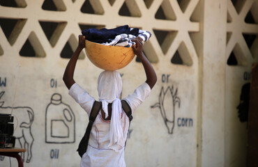horizontal  photpgraphy: closeup of a skinny African girl in white clothes and head wrap, carrying a full yellow bowl on her head, by the white wall- , outdoors on a sunny day, in the Gambia, Africa