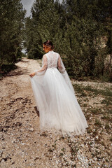 view from back. woman in wedding dress walks along mountain path between trees.