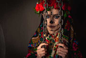 young serious girl with makeup for the Day of the Dead with a wreath of flowers on his head, in a mesh with flowers handkerchief holds a metal chain and looks through them. Halloween or La Calavera