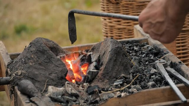 Slow motion: professional blacksmith hand heating metal piece in diy outdoor forge at summer historical medival festival - close up. Craftsmanship, reenactment, handwork, revival concept