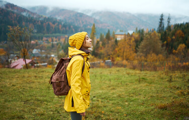 Side view tourist woman in yellow wear standing on meadow with beautiful mountains view. Backpacking girl looking forward and getting ready to hiking