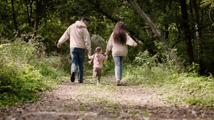Mom, dad, daughter are playing in park, run along path holding hands. Happy child is holding hands of parents. Happy family walks in forest holding hands in spring, summer. Teamwork. Family weekend
