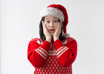 A teenage girl in a Santa Claus hat and a red Christmas sweater is surprised and holds her hands on her cheeks. The concept of Christmas and New Year