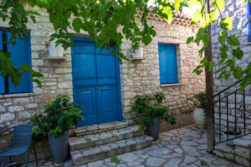 Fototapeta na wymiar Sun shining through green tree branches in summer in Greek traditional yard with stone walls and blue doors and windows shutters. Summer travel locations architecture