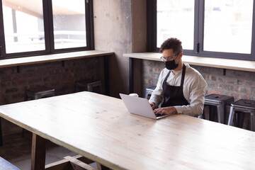 African american woman wearing apron and face mask using laptop at gin distillery