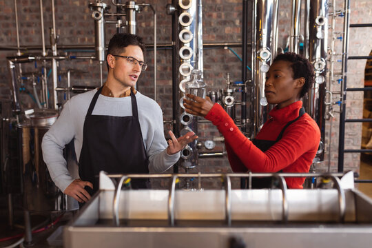 Caucasian man and african american woman checking bottle of gin in glass at gin distillery