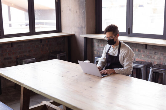 African american woman wearing apron and face mask using laptop at gin distillery - Powered by Adobe