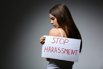Young woman holding paper with text STOP HARASSMENT on grey background