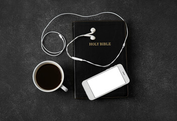 Holy Bible, earphones, mobile phone and cup of coffee on dark background