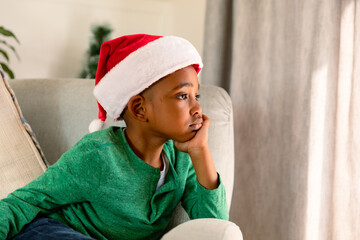 Thoughtful african american boy wearing santa hat and sitting in armchair at christmas time