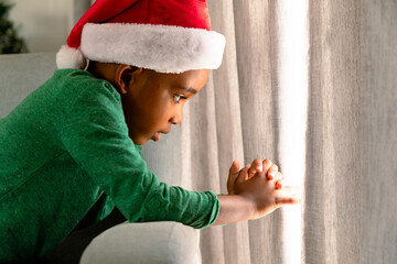 African american boy wearing santa hat and looking through window at christmas time