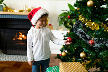 Happy caucasian boy wearing santa hat, decorating christmas tree at christmas time