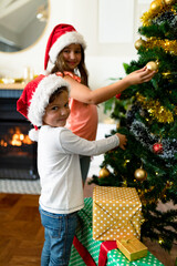 Happy caucasian sibling wearing santa hats, decorating christmas tree
