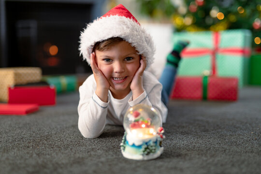 Happpy caucasian boy wearing santa hat, looking at snow globe at christmas time