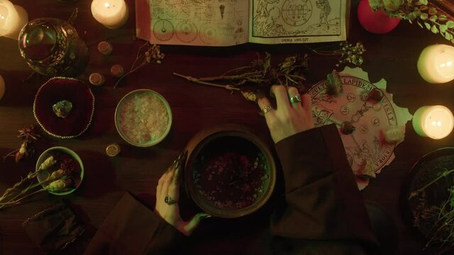 Witch cooking potion elixir top view. Table with spellbook for making rite. Herbs, burning candles, crystal magic ball for sacrifice. Halloween theme, full moon, witchcraft. 