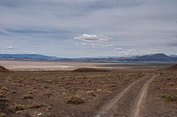 High Desert, Nevada, USA - May 17, 2011: Wide landscape with vehicle trail through dirt and dry lake under blue-gray cloudscape. Snow covered mountain range in back. Shrubs in front.