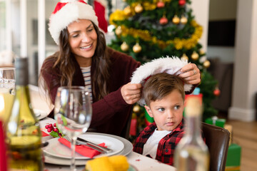 Caucasian mother putting on santa hat on her son head at christmas table