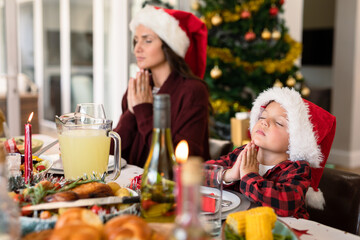 Caucasian mother and son wearing santa hats praying at christmas table