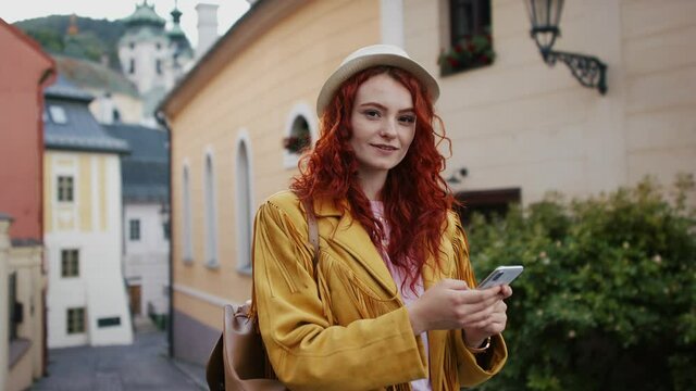 Young woman outdoors on trip in town, using online map on smartphone and looking at camera.