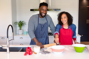Happy african american couple wearing aprons, baking together and looking at camera