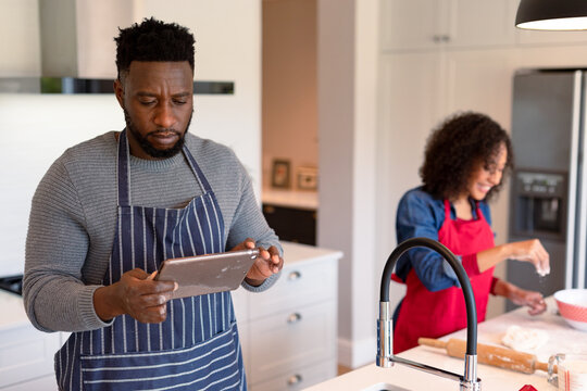 Focused african american couple wearing aprons, baking together and using tablet